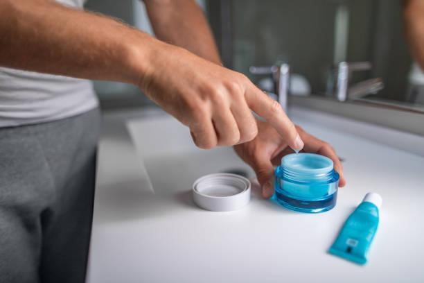 mens die de huidverzorgingsregime van de roomochtend toepast hydrateert gezichtshuid bij badkamerspiegel. mannelijke schoonheidsmensen huidverzorging close-up van product pot en vinger putting lotion - skin condition creams stockfoto's en -beelden