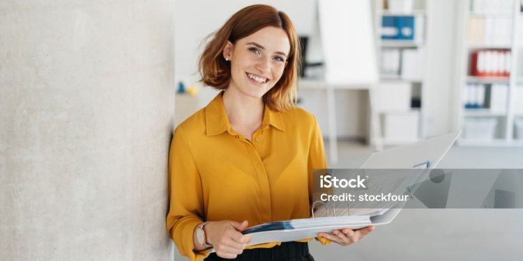 Attractive young office worker holding a large open binder as she looks at the camera with a sweet friendly smile